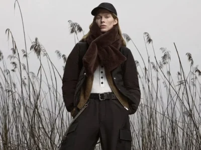 Model in black outerwear and cap stands among tall reeds, wearing a large brown scarf and belted cargo pants
