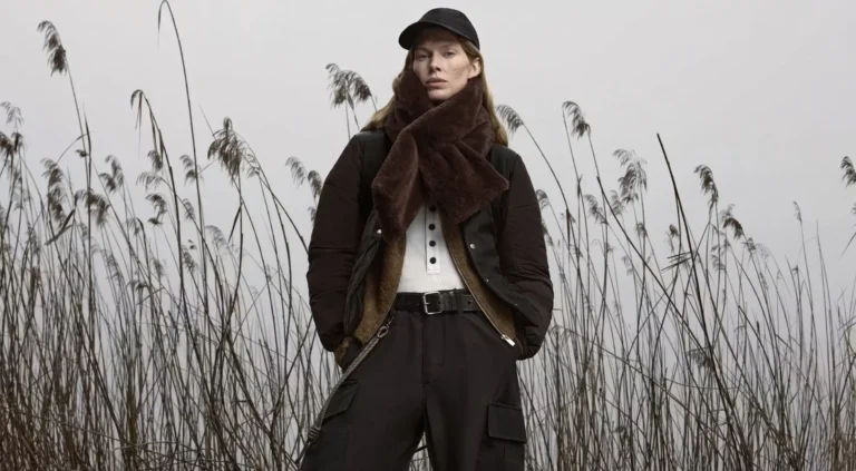 Model in black outerwear and cap stands among tall reeds, wearing a large brown scarf and belted cargo pants