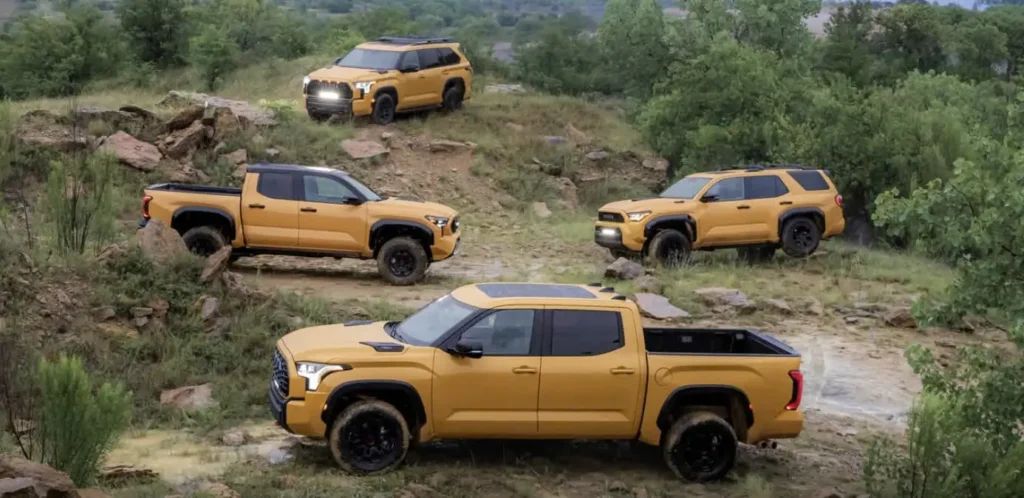 Four mustard-yellow pickup trucks and SUVs navigate a rocky off-road trail surrounded by green trees and hills