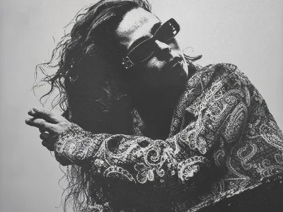 Lov'Nee poses in sunglasses and a paisley shirt, leaning back against a plain light background