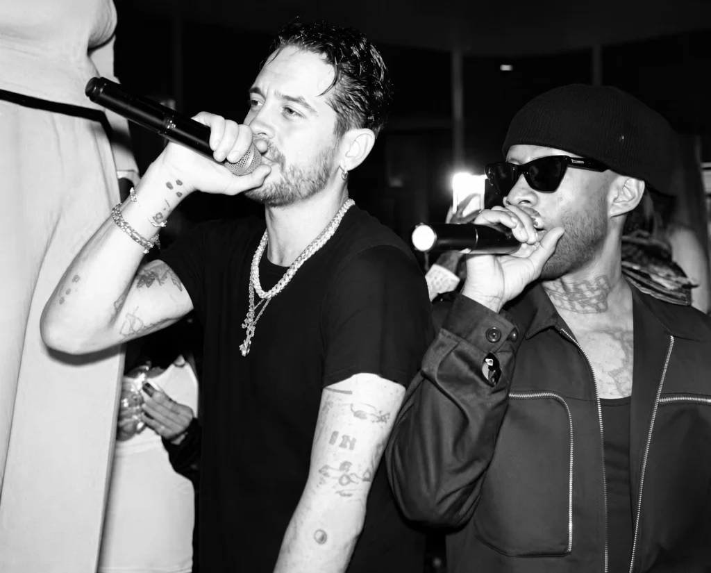 G-Eazy and Ty Dolla $ign perform into microphones, wearing black outfits, jewelry, sunglasses and a beanie in a dim venue
