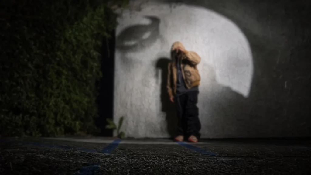 Person in a tan jacket and black pants stands before a large white abstract mural, with greenery at left