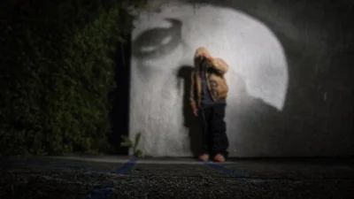Person in a tan jacket and black pants stands before a large white abstract mural, with greenery at left