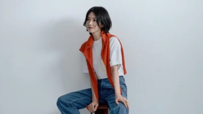 Song Hye Kyo seated on a stool against a white wall, wearing a white T-shirt, blue jeans, and an orange sweater draped over the