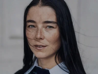 Close-up portrait of Julie Kegels with long dark hair, wearing a pale blue collared shirt against a gray background