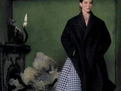 Woman in a black cape and blue-and-white checked skirt stands beside a black cat sculpture and candle in a dark green room