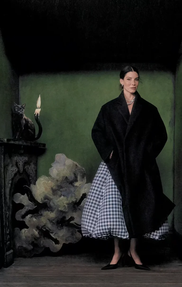 Woman in a black cape and blue-and-white checked skirt stands beside a black cat sculpture and candle in a dark green room