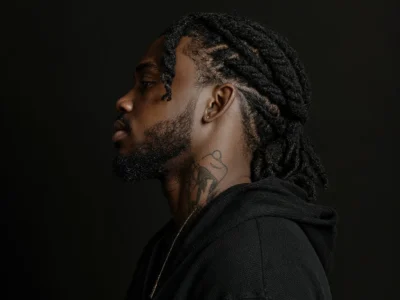 Profile of Che Ecru with braided hair, beard, neck tattoo, and black hoodie against a dark background