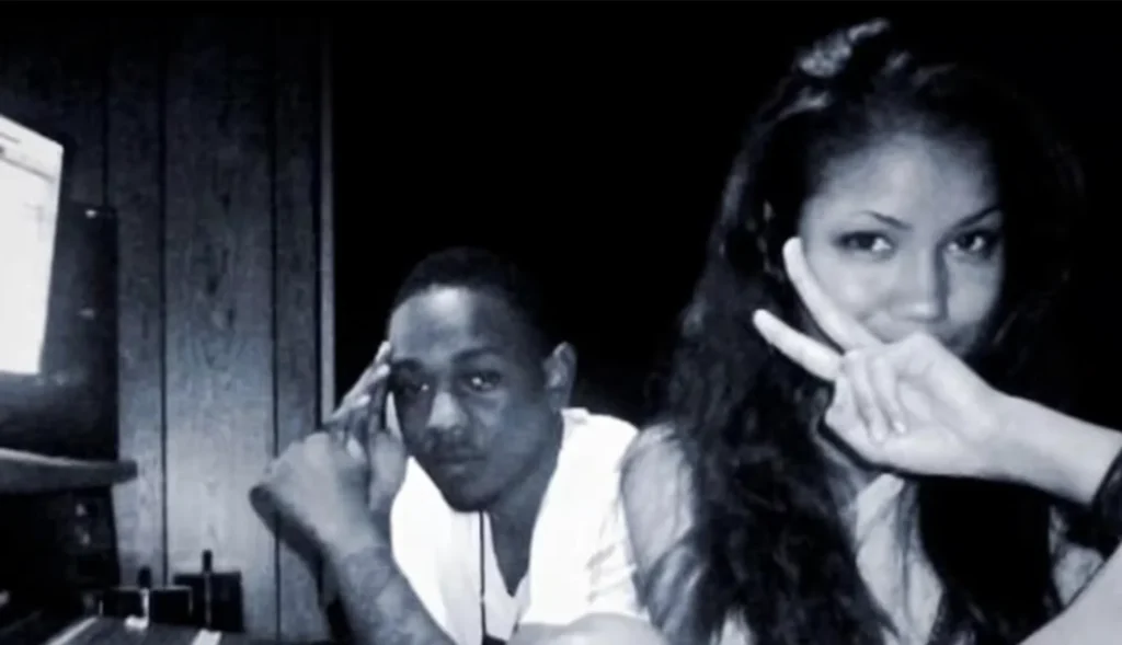 Jhené Aiko and Kendrick Lamar pose in a dim room, each holding two fingers near their eyes in a black-and-white image