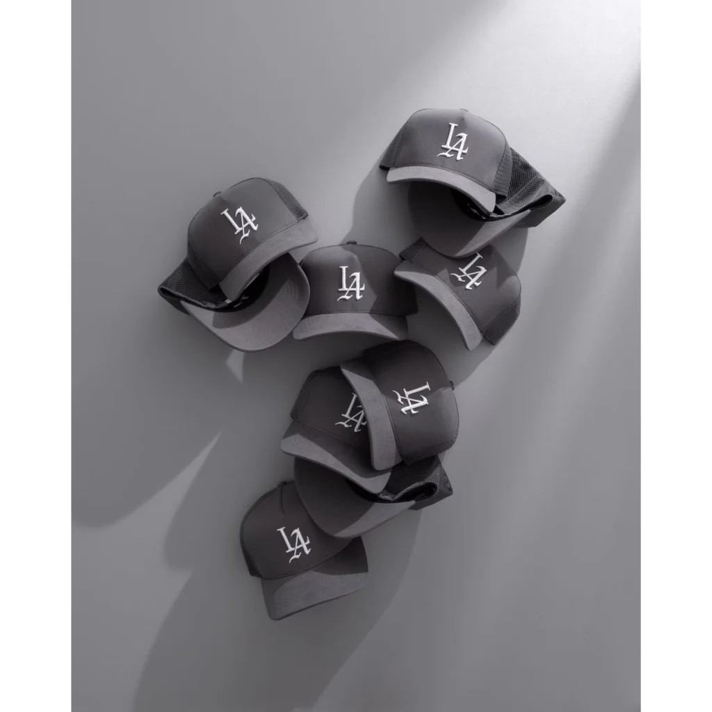 Five charcoal mesh-back trucker caps with white LA logos arranged on a gray wall with strong shadows