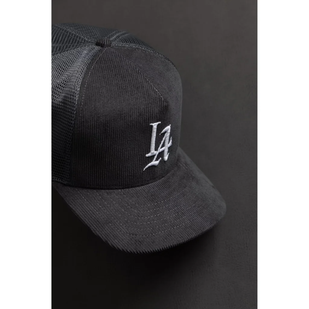 Black corduroy trucker cap with mesh back and white embroidered logo, angled on a dark background