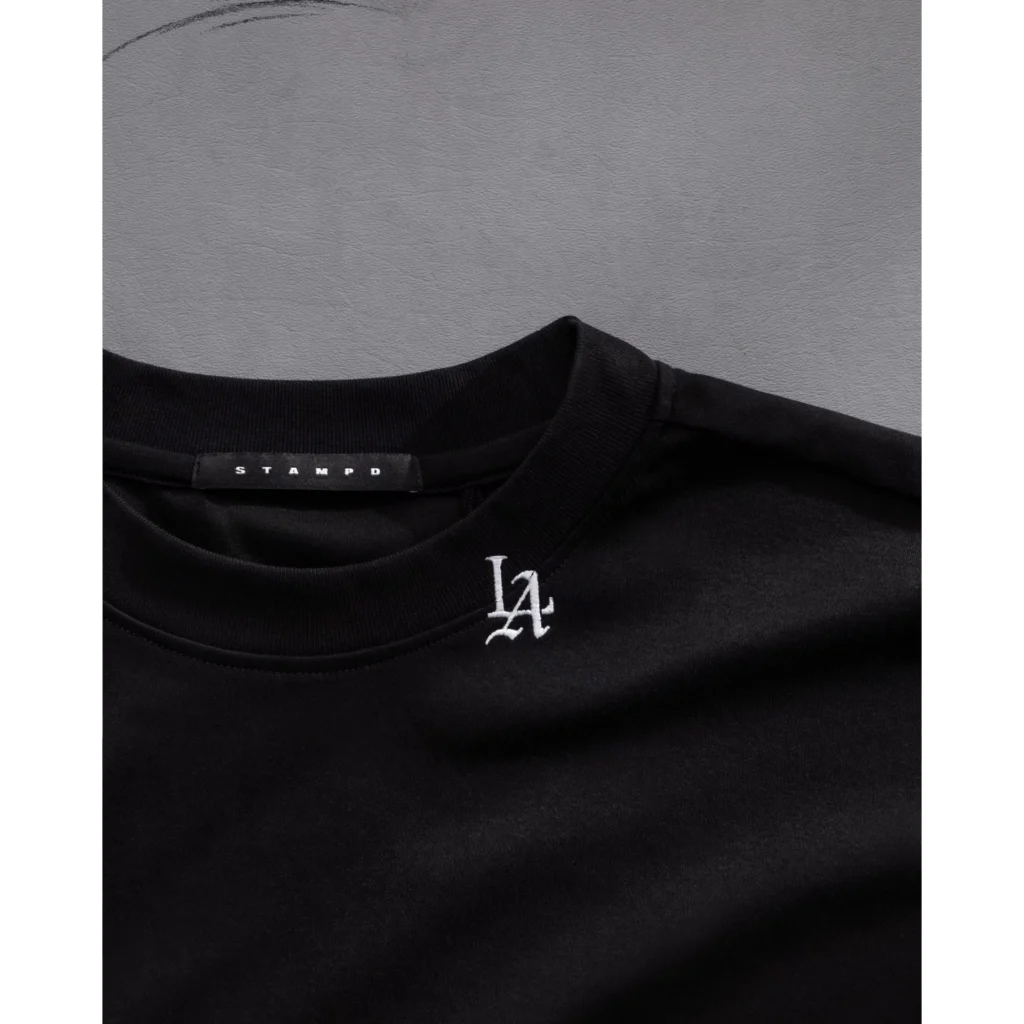 Close-up of a black Tee with ribbed crew neck, Stampd neck label, and small white embroidered chest logo against gray