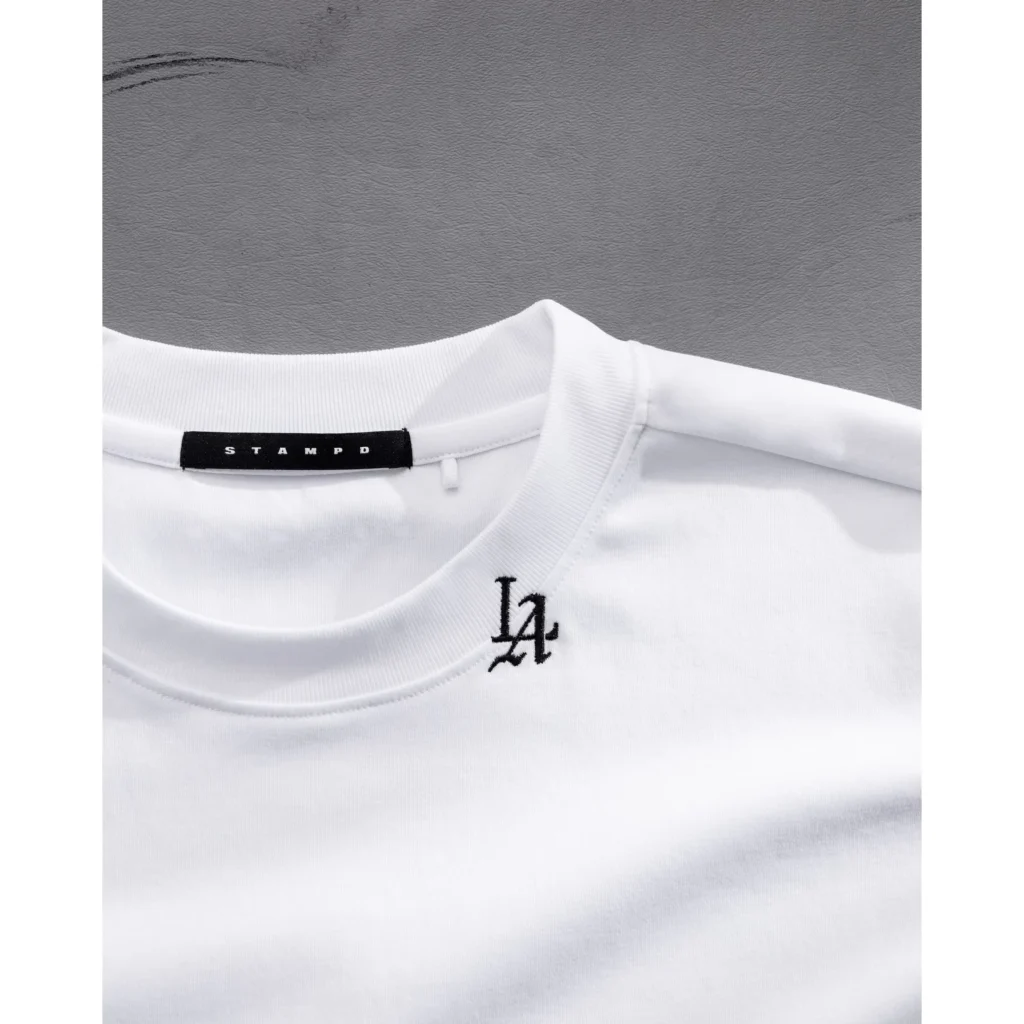 Close-up of a white relaxed-fit T-shirt with ribbed collar, black LA embroidery, and Stampd neck label against gray fabric