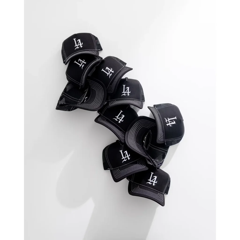 Nine black mesh trucker caps with white LA logos and contrasting stitching arranged in a vertical pile on white background