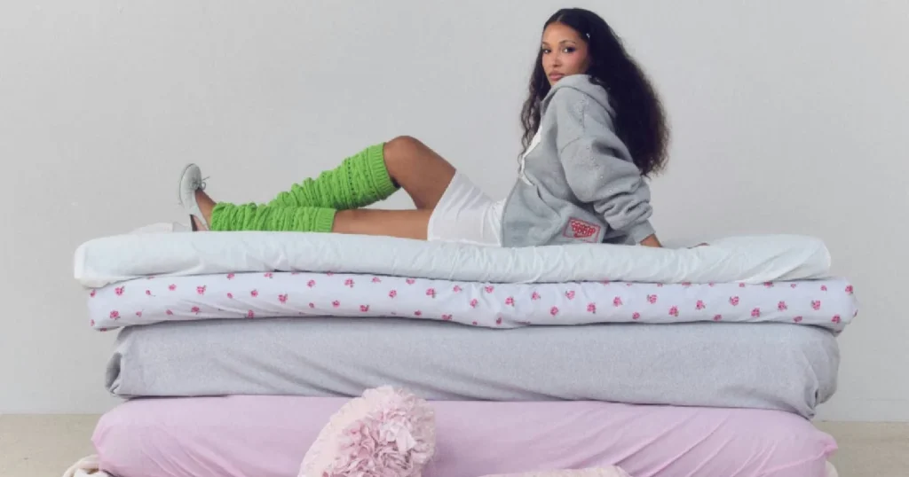 Yasmina reclines on stacked mattresses wearing a gray hoodie, white shorts, bright green leg warmers, and white sneakers