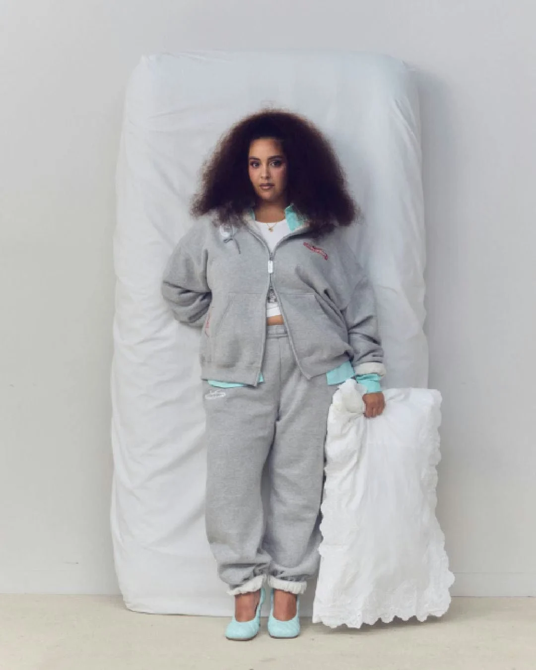 Yasmina stands beside a bed in a gray tracksuit with turquoise accents, holding a white pillow and wearing turquoise shoes