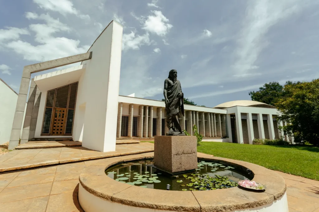 White modernist building and bearded statue beside a lily pond under a blue, cloud-filled sky