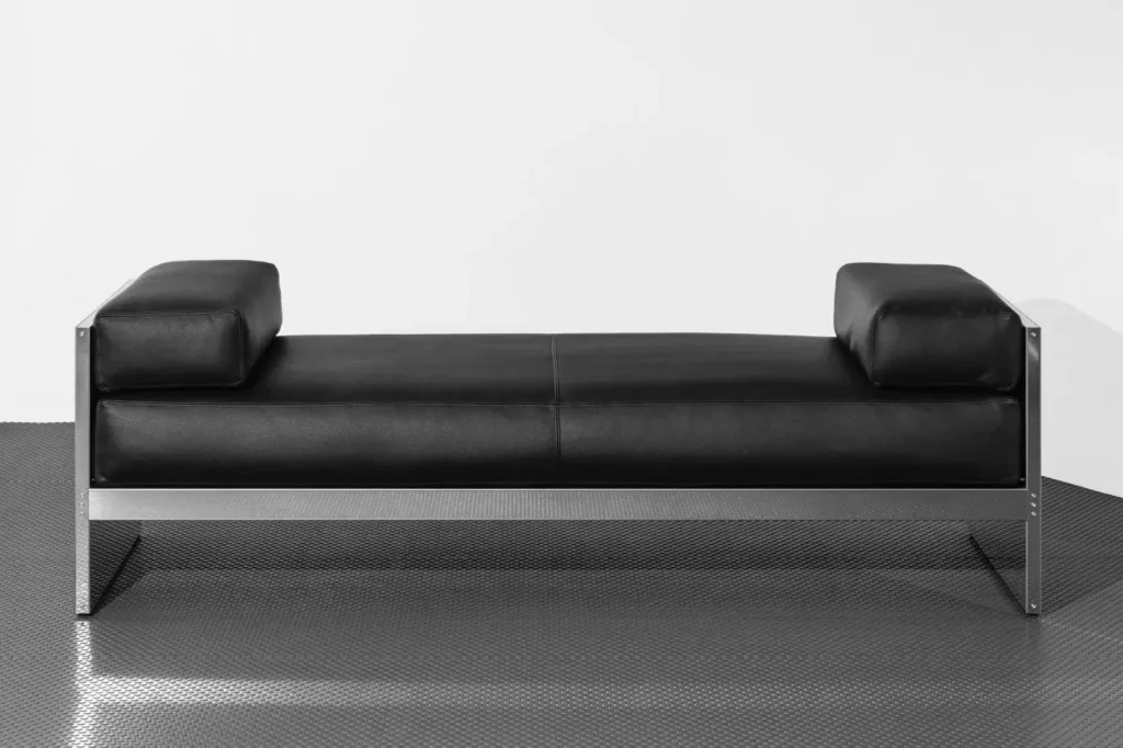 Black leather sofa with square armrests and a brushed metal frame on a textured metal floor against a white wall