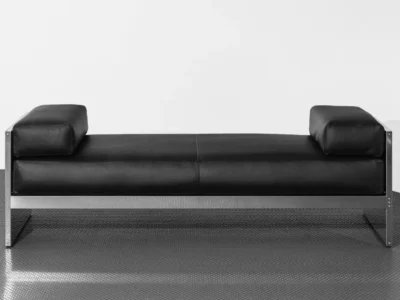 Black leather sofa with square armrests and a brushed metal frame on a textured metal floor against a white wall