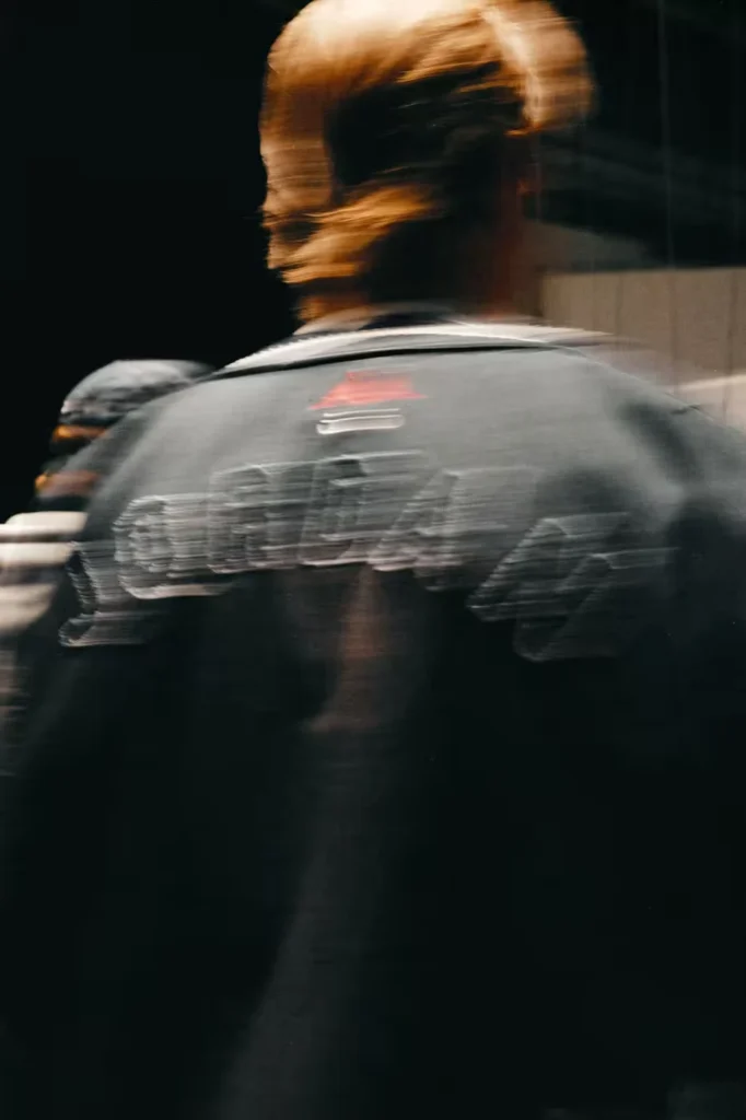Blurred figure seen from behind in a gray graphic T-shirt, with long blond hair, against a dark background