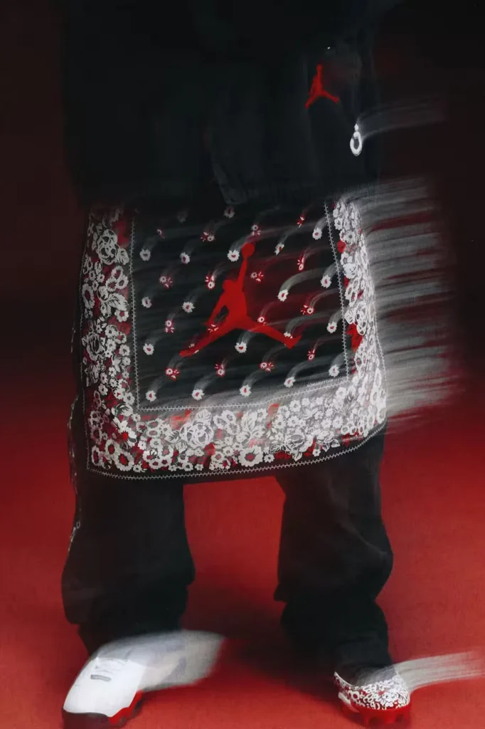 Person wearing a black floral-patterned apron with a red Jumpman logo, black pants and white cleats against a red background