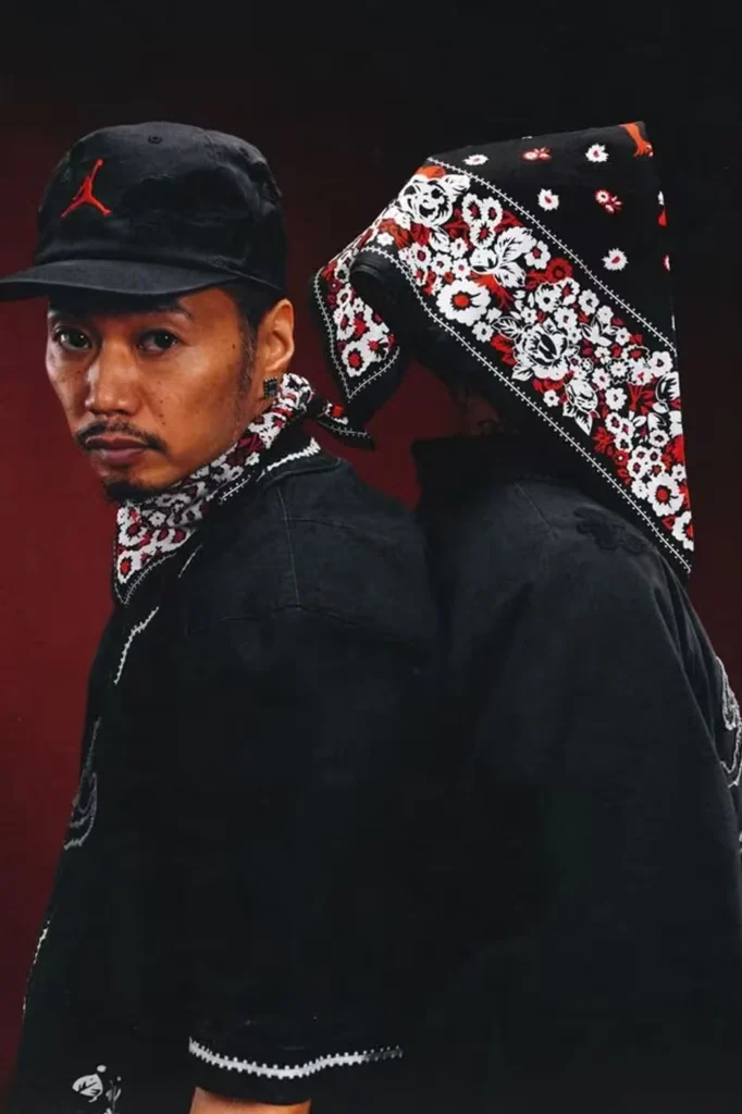 Two people in black clothing and floral bandanas pose back-to-back against a dark red background; one wears a Jordan cap