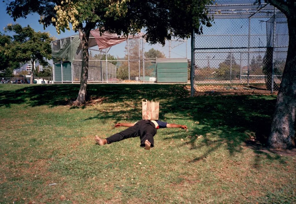 Person lying spread-eagle on grass with a paper bag over their head, wearing black pants, beside fenced courts and trees