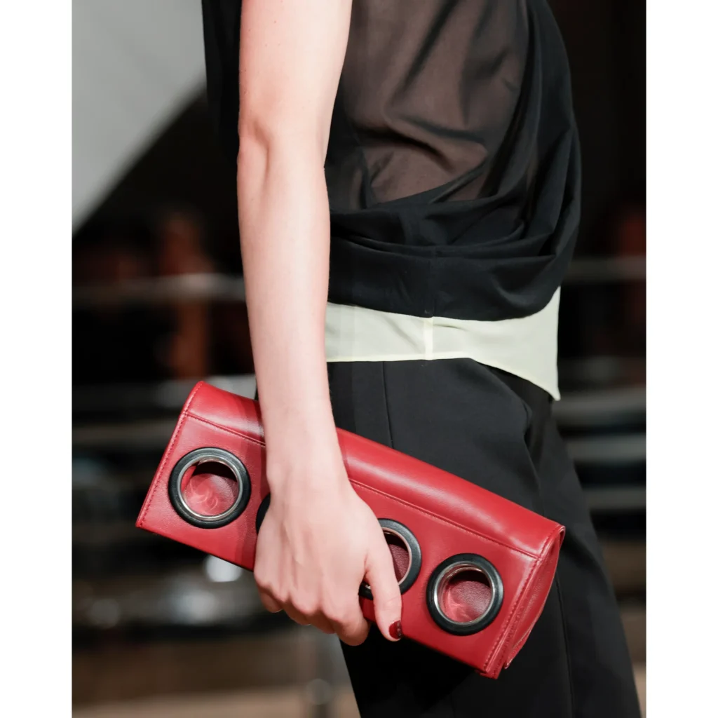 Cropped model in a black layered outfit holding a red leather clutch with three large black-rimmed grommets