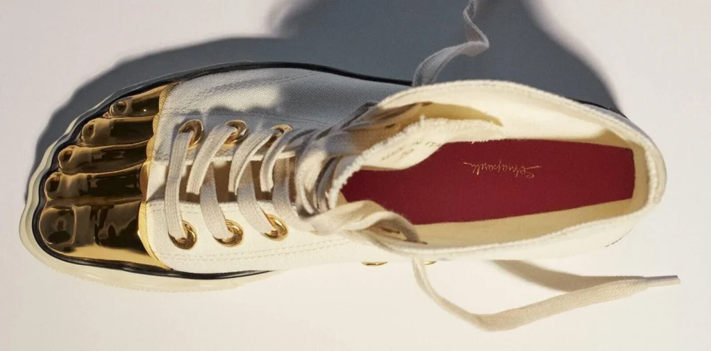 White low-profile shoe with gold metal toe detailing, cream laces, and a burgundy insole on a pale surface