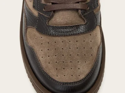 Close-up of a brown suede sneaker with dark textured leather panels, brown laces, perforated toe, and thick rubber sole