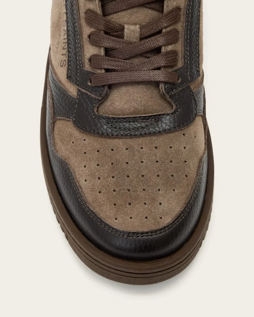 Close-up of a brown suede sneaker with dark textured leather panels, brown laces, perforated toe, and thick rubber sole