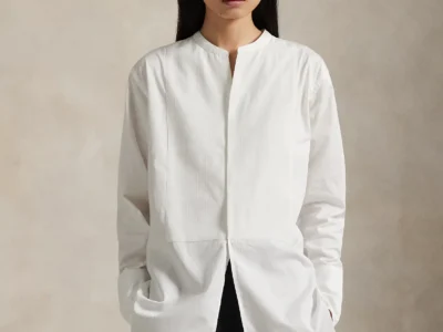 Model wearing a white collarless button-front tunic with curved hem and black trousers, hands in pockets against a beige