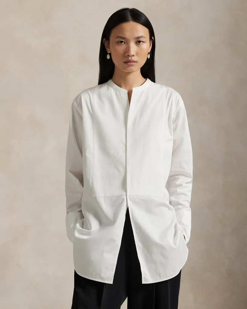 Model wearing a white collarless button-front tunic with curved hem and black trousers, hands in pockets against a beige