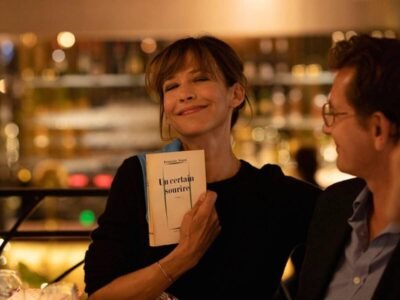 Sophie Marceau smiles while holding a French book at a warmly lit restaurant table with two people beside her