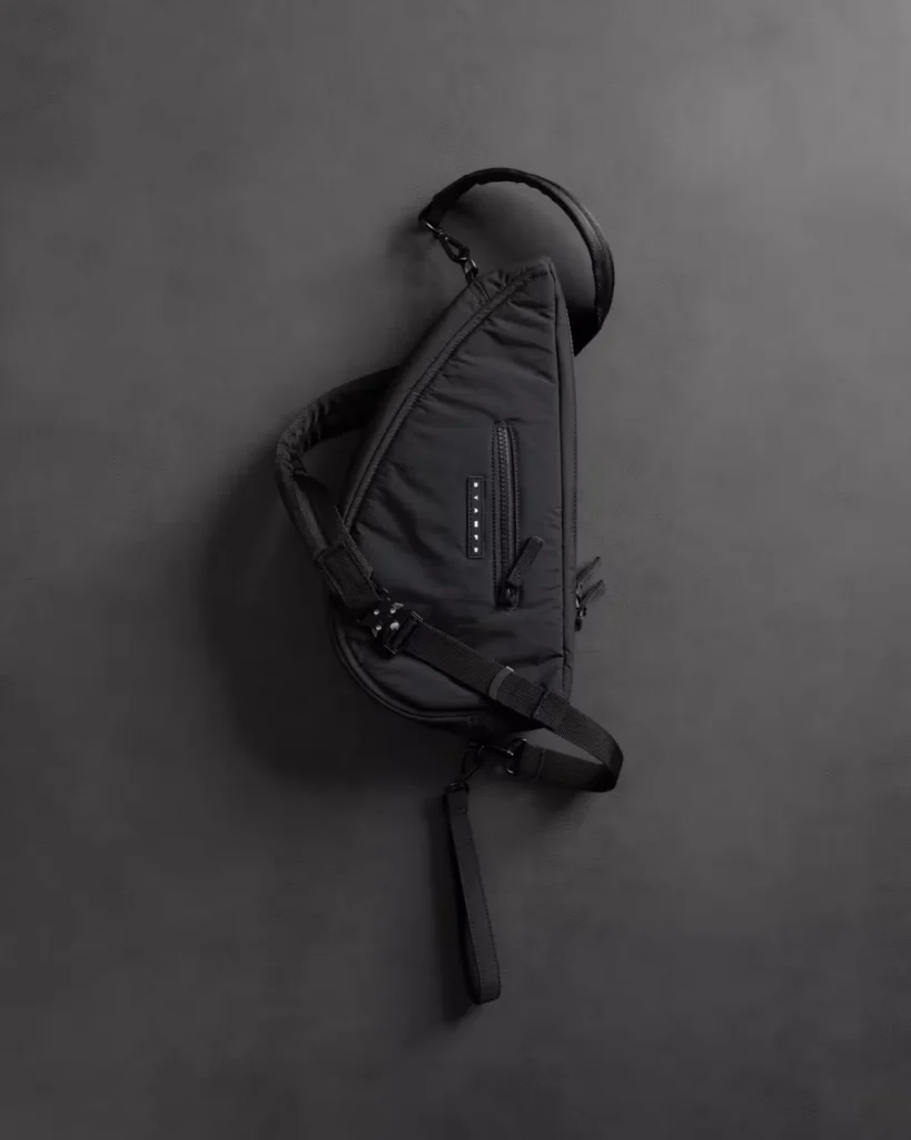 Black sling shoulder bag with adjustable webbing strap and front zip pocket, displayed against a dark gray background