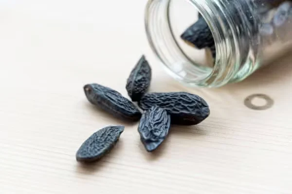 Black tonka beans scatter on a pale wooden surface beside a tipped glass jar