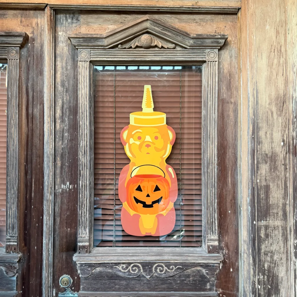 Bright yellow honey bear cutout holding an orange jack-o’-lantern, displayed behind a window with wooden shutters