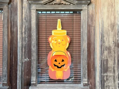 Bright yellow honey bear cutout holding an orange jack-o’-lantern, displayed behind a window with wooden shutters