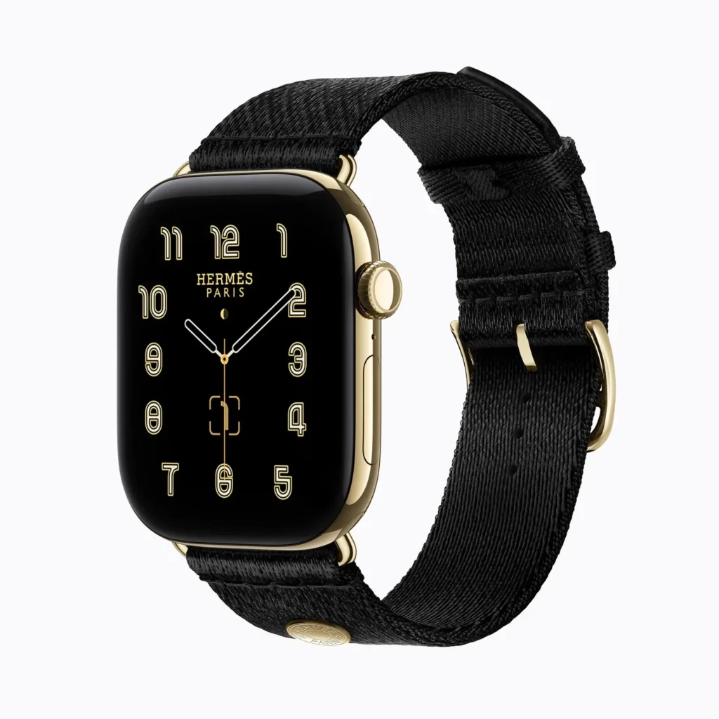 Hermès Apple Watch with black woven band, gold-toned case, and black face showing gold numerals on white background