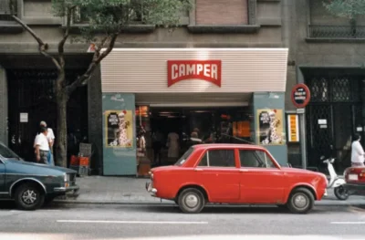 Camper storefront with a red sign, pictured behind a parked vintage red sedan on a city street