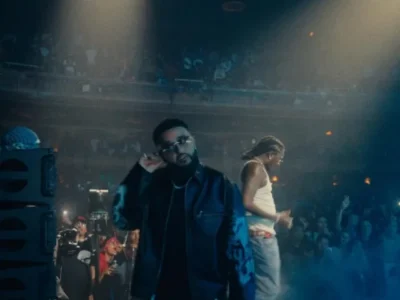 Two performers on a smoky concert stage, one in a black jacket and glasses, another in a white tank top behind him