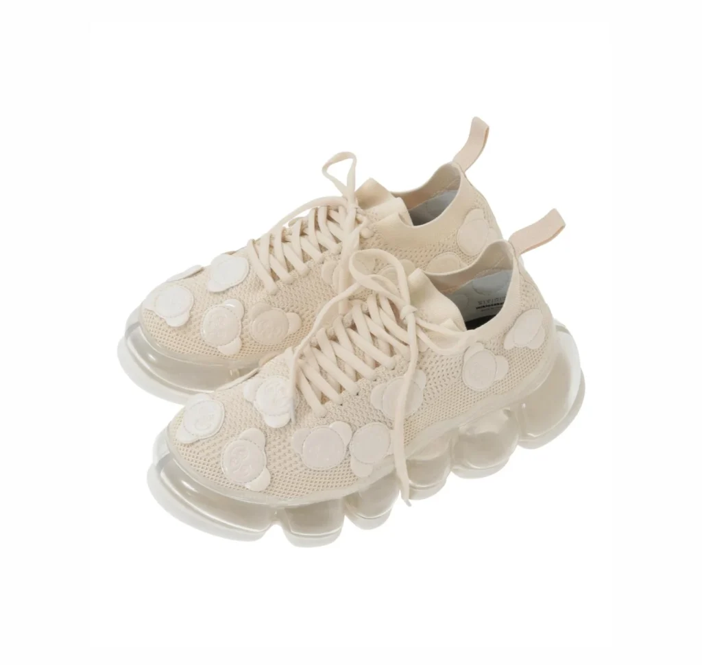 Pair of cream knit sneakers with circular patches and translucent, segmented soles on a white background
