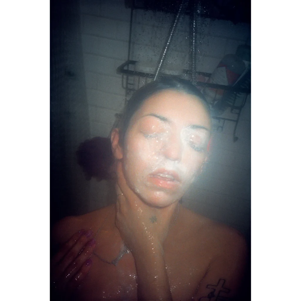 Close-up of a person with shaved hair, glitter on their face and body, and a hand with long purple nails in a dim room