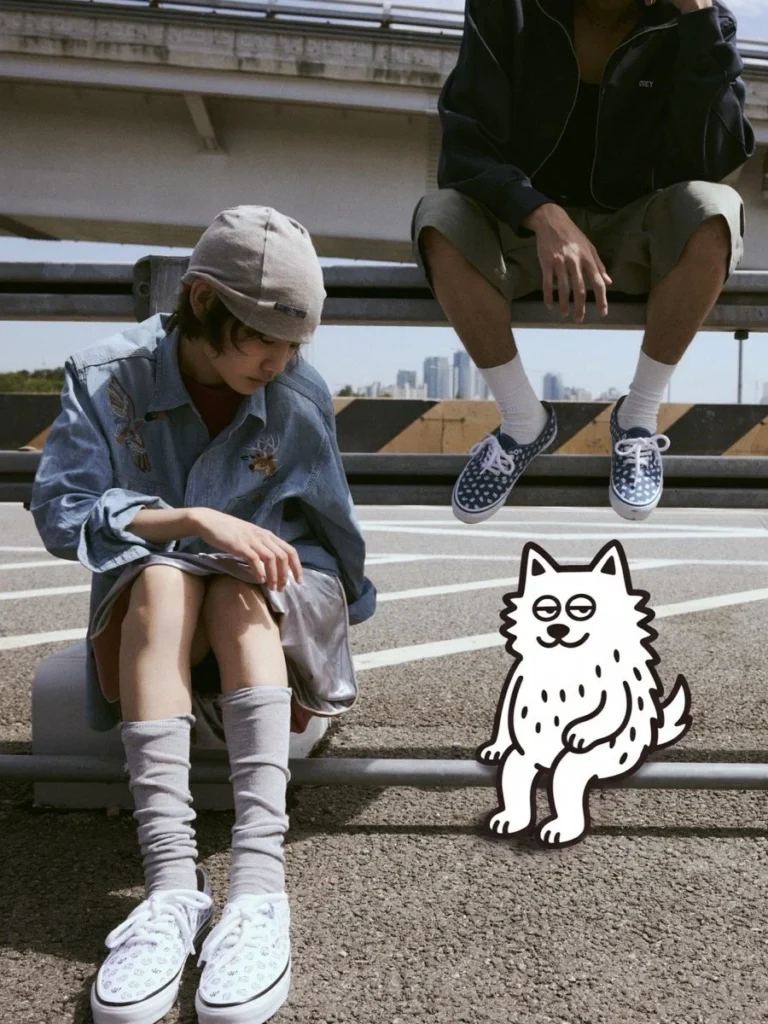 Two people pose on an urban overpass, one seated in embroidered denim and one perched above, beside a cartoon cat sticker
