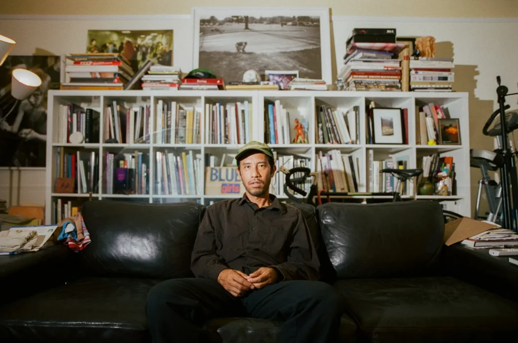 Jerry Hsu sits on a black leather sofa wearing a dark shirt and green cap, with bookshelves behind him