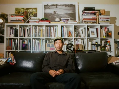 Jerry Hsu sits on a black leather sofa wearing a dark shirt and green cap, with bookshelves behind him