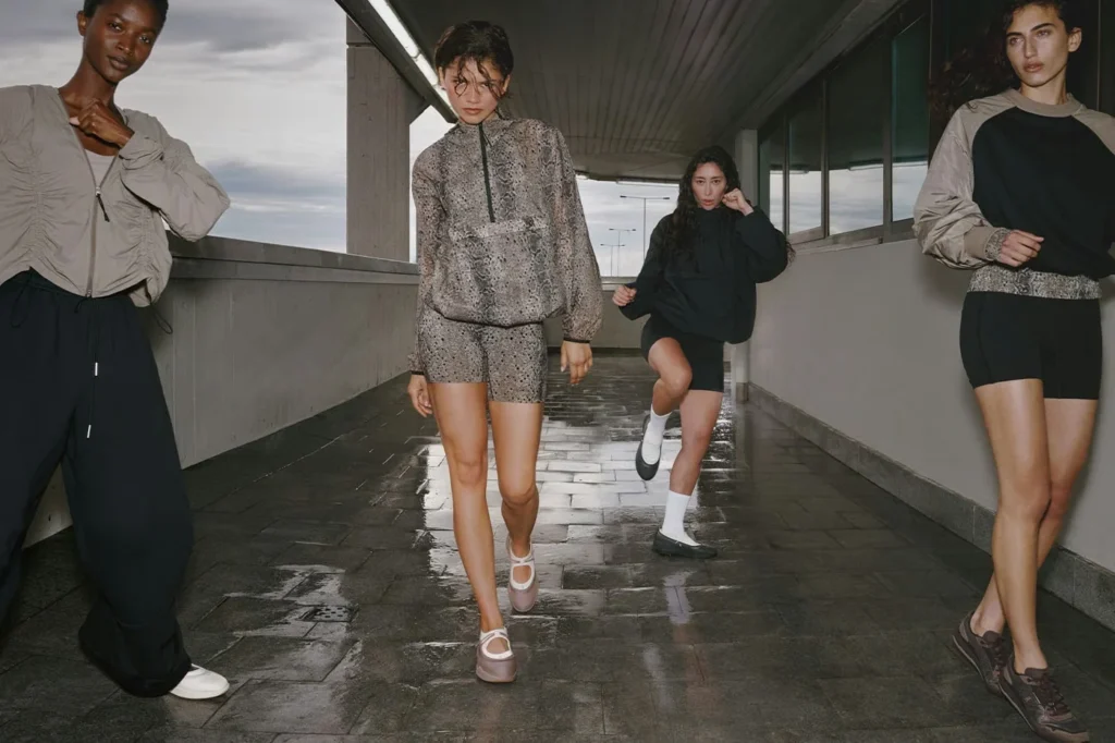 Four models walk through a wet concrete passage, wearing gray, black, and snakeskin athletic clothing and sneakers