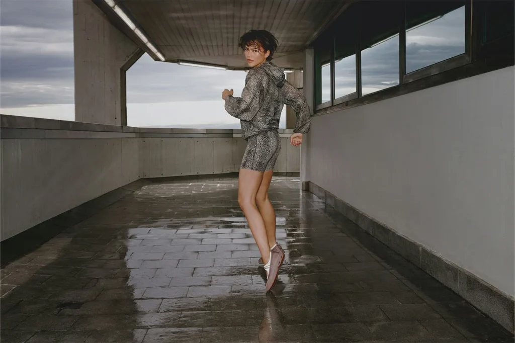 Zendaya in a gray patterned hooded jacket and shorts poses mid-stride in a wet, windowed concrete corridor