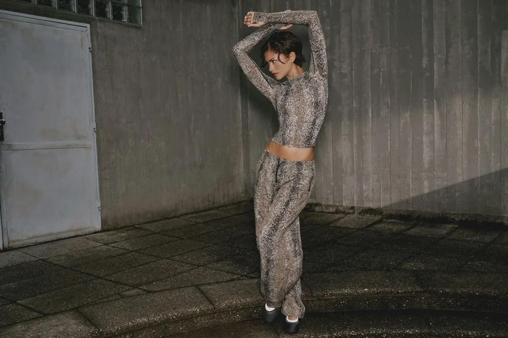 Zendaya poses with arms raised in a snakeskin-print long-sleeve crop top and matching pants by a concrete wall