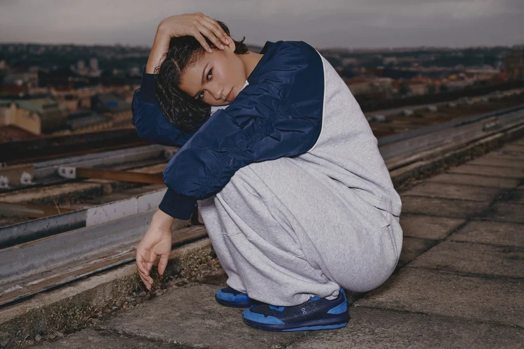 Zendaya crouches on a rooftop in a navy jacket, gray pants and blue sneakers, with a cityscape behind her